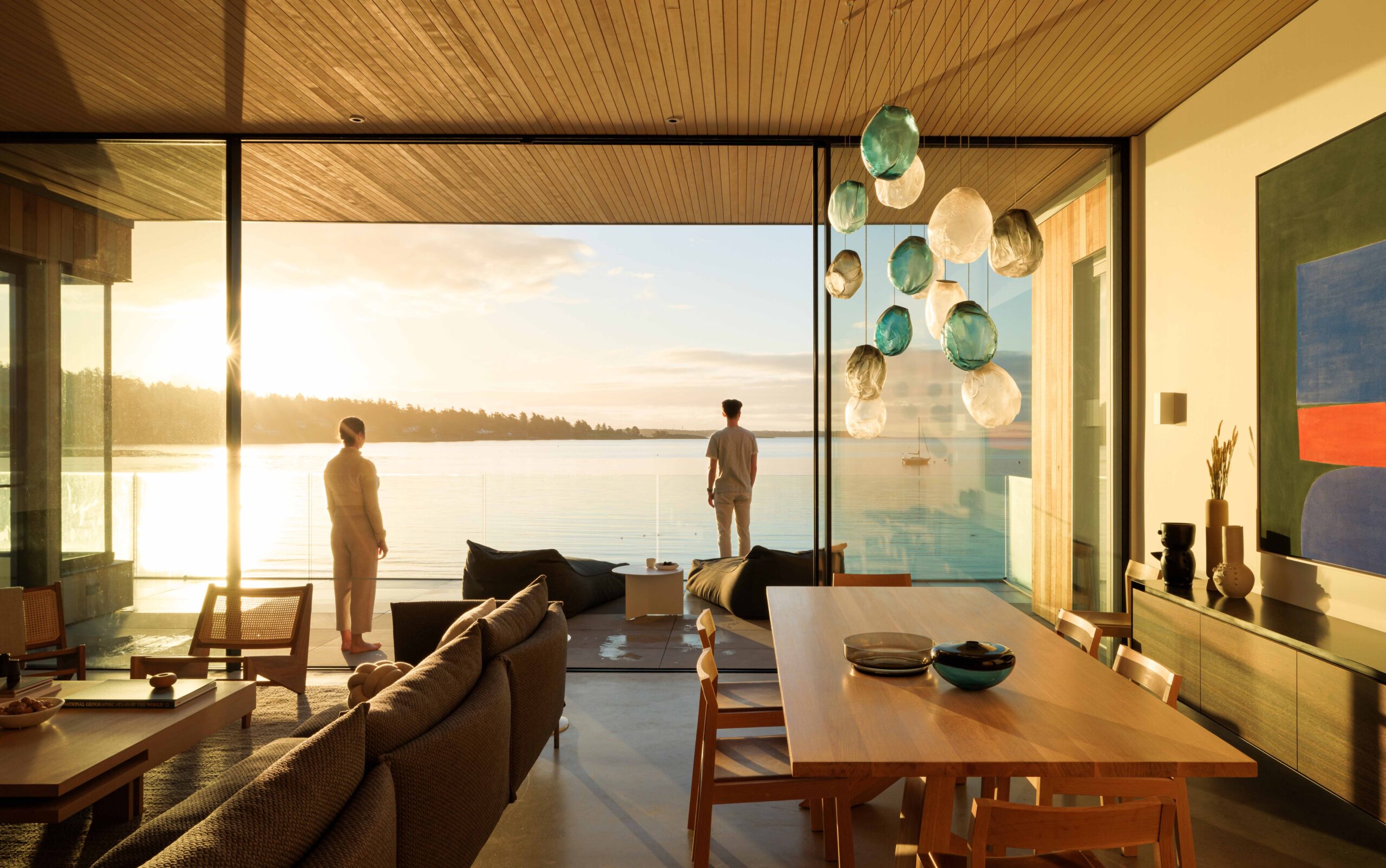 A man and woman watch the sunrise over a water from a deck with large open sliding doors. In the foreground a light oak dining table and chairs on the right has a blue white and grey hand blown glass light fixture above and on the left a modern sofa with textured grey fabric is grouped with a wood and cane-backed arm chair. The the Falken Reynolds designed interior and view are peaceful and calming. A modernist painting is hung on the right wall.